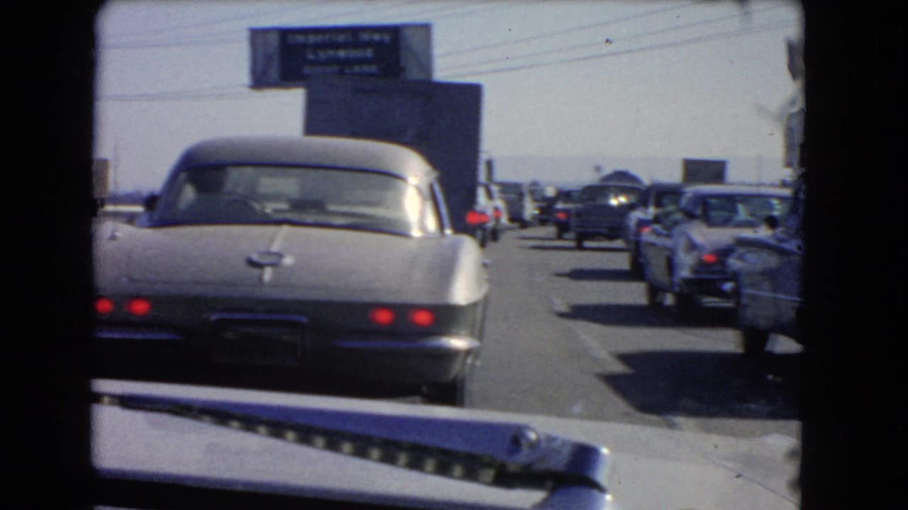 1966 California freeway traffic seen from inside a car, with a silver sports car ahead and an Imperial Highway Lynwood right-lane sign overhead.