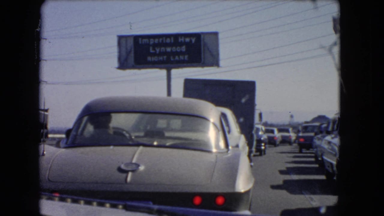 A silver sports car sits ahead in slow traffic near an Imperial Highway Lynwood right-lane sign.