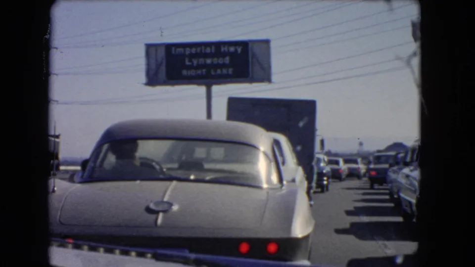 In-car view of 1960s California freeway congestion, with a silver sports car centered and multiple vehicles lined up beside it.