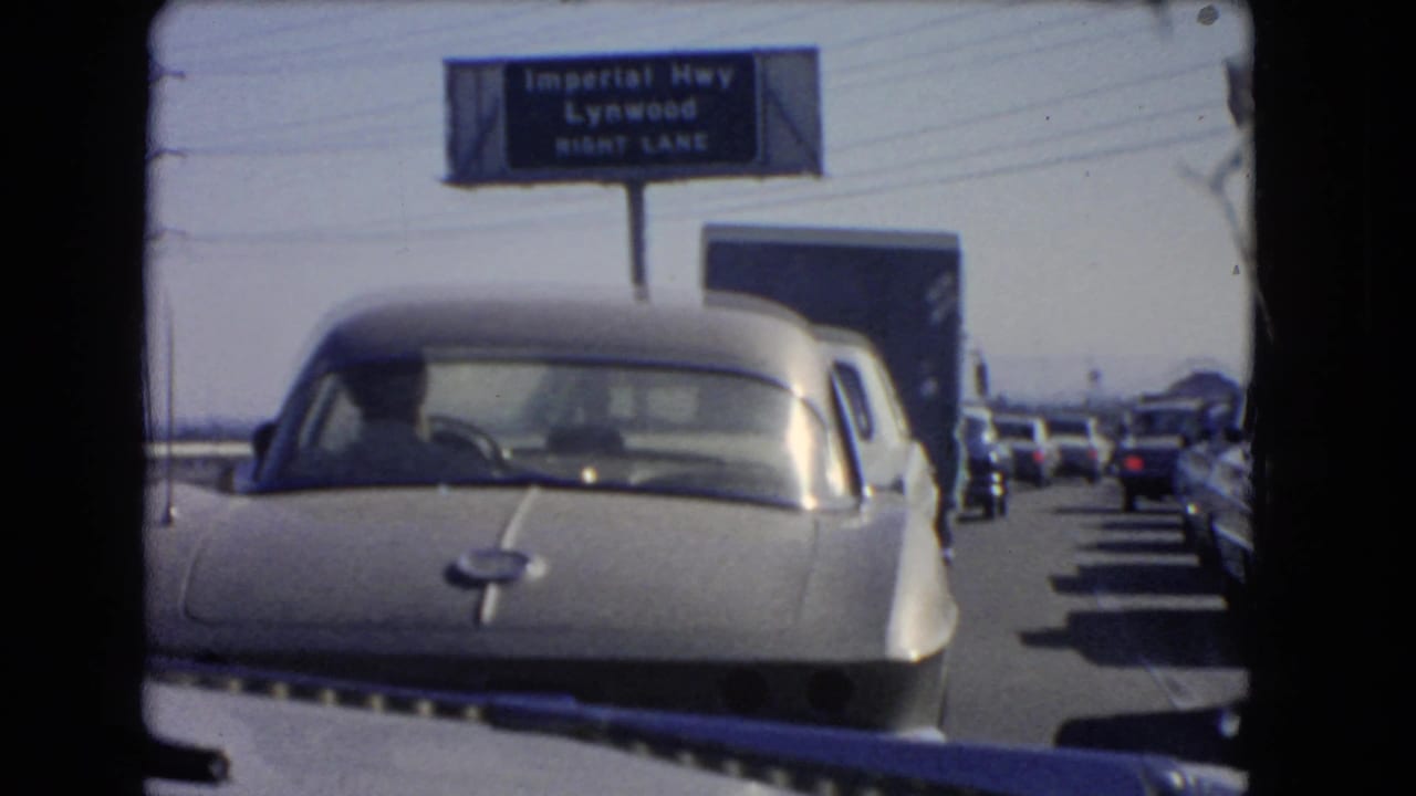 A silver sports car fills the foreground during a 1966 California freeway standstill, with the Imperial Highway Lynwood sign visible above.