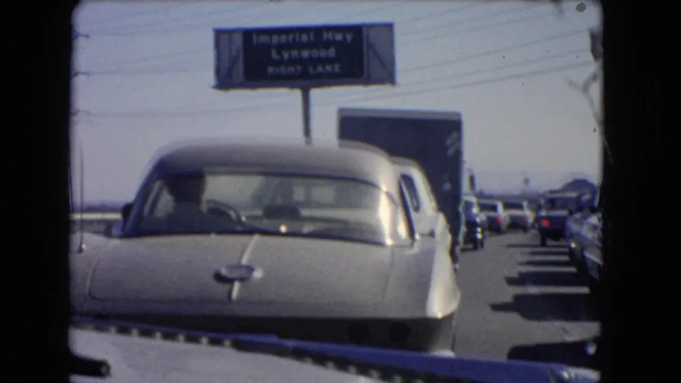 A silver sports car fills the foreground during a 1966 California freeway standstill, with the Imperial Highway Lynwood sign visible above.