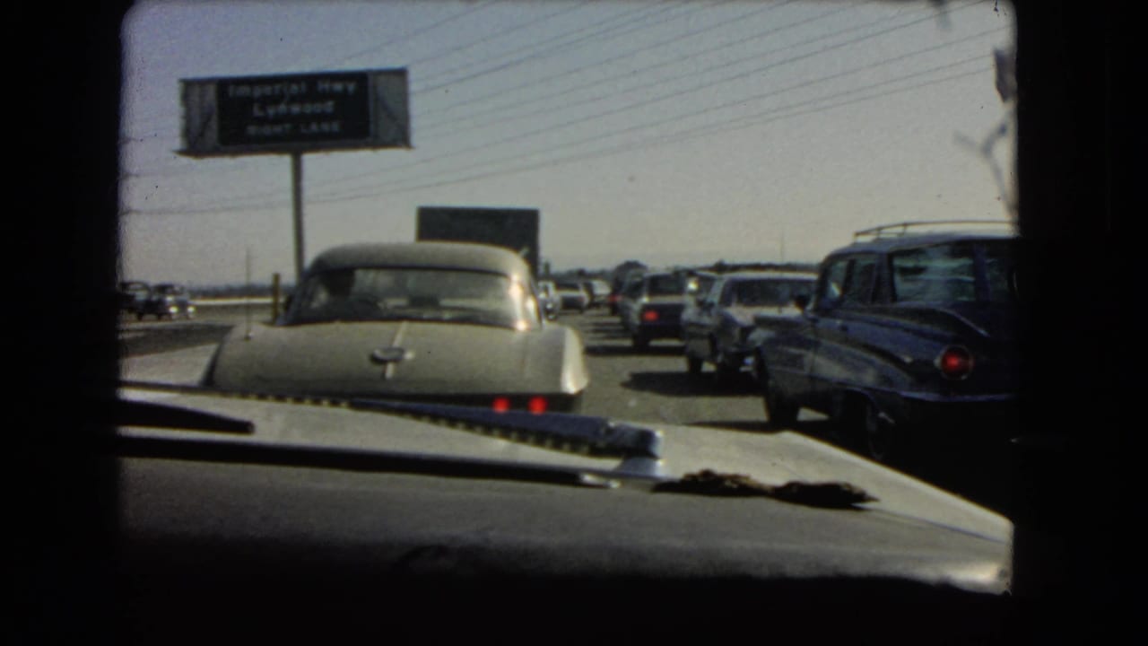 Wider in-car view of 1966 California freeway congestion, showing multiple lanes of classic cars and the Imperial Highway Lynwood sign in the distance.