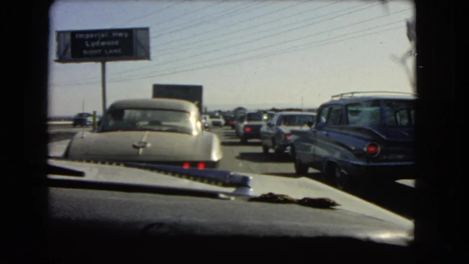 In-car 1966 California road view showing a line of older vehicles, including a station wagon, beneath overhead signage.