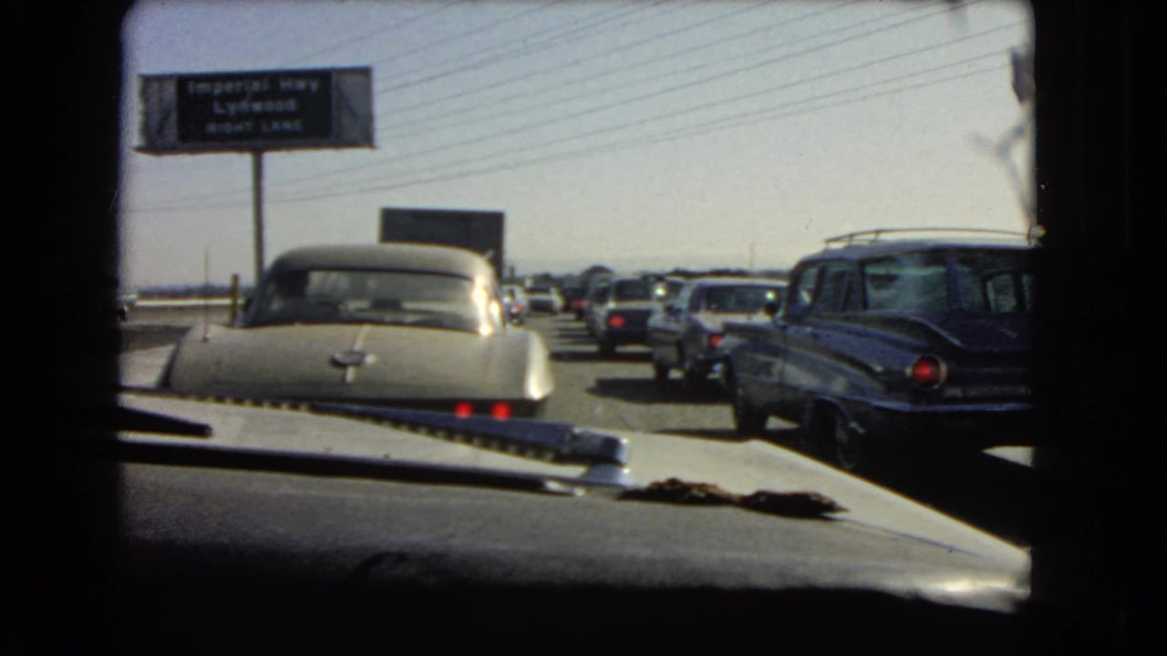 1960s California home-movie frame of traffic congestion filmed through the windshield, with overhead signage and classic cars ahead.