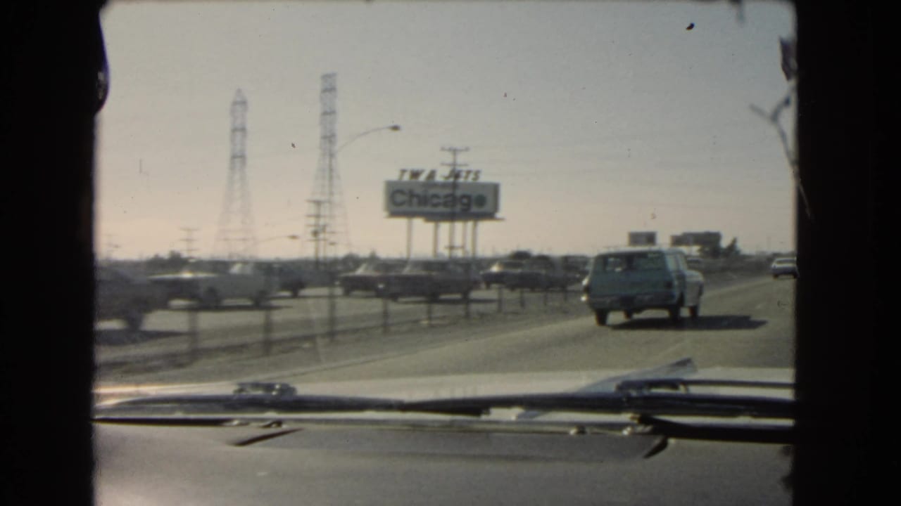 Mid-1960s 8mm windshield shot in California showing a station wagon ahead, roadside traffic, transmission towers, and a TWA Jets Chicago billboard.
