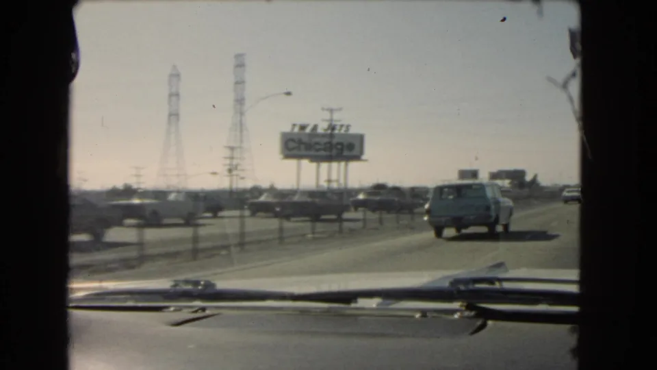 Mid-1960s 8mm windshield shot in California showing a station wagon ahead, roadside traffic, transmission towers, and a TWA Jets Chicago billboard.