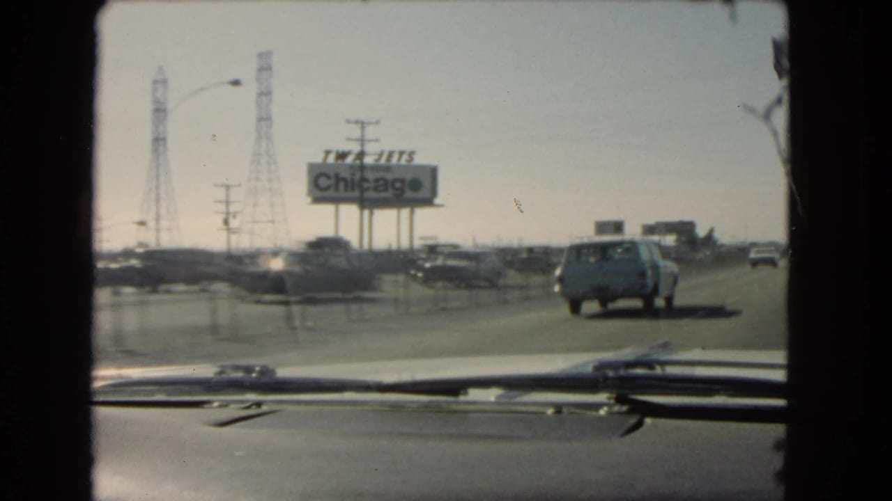1966 California home-movie frame from inside a car, with a station wagon ahead, passing highway traffic, power towers, and a TWA Jets Chicago sign.