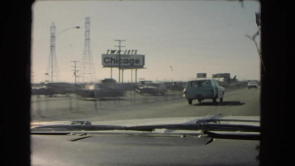 1966 California home-movie frame from inside a car, with a station wagon ahead, passing highway traffic, power towers, and a TWA Jets Chicago sign.