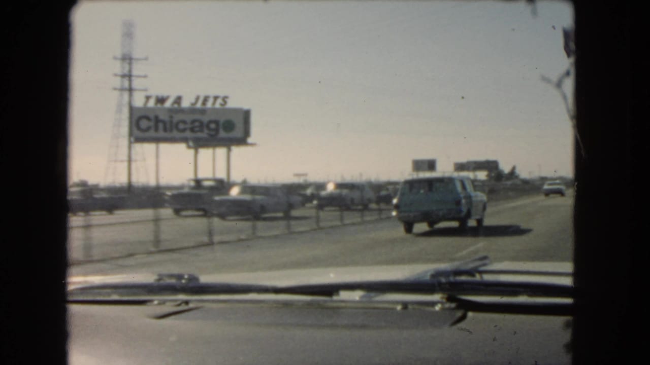 Windshield view on a 1966 California highway, showing a blue station wagon, passing cars, utility towers, and a roadside TWA Jets Chicago sign.