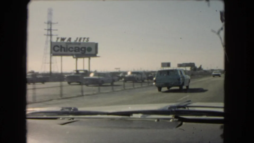 Windshield view on a 1966 California highway, showing a blue station wagon, passing cars, utility towers, and a roadside TWA Jets Chicago sign.