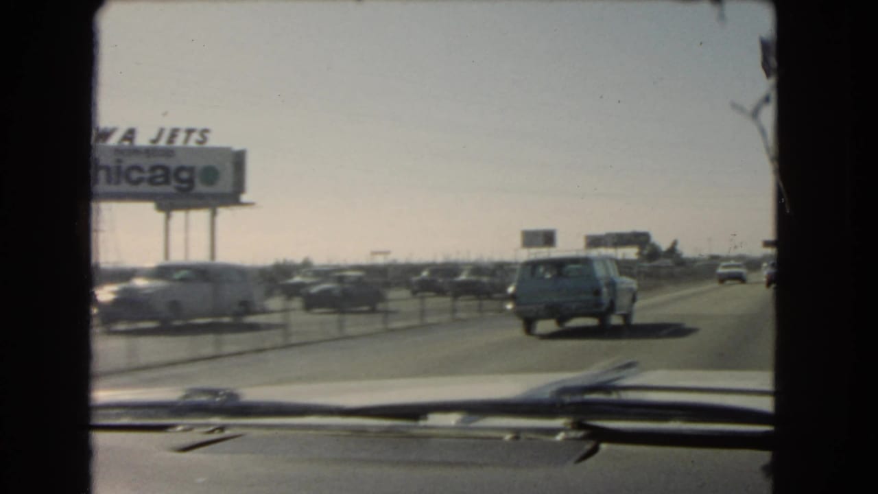 From inside the car, the 1966 California highway scene shows motion-blurred traffic, a cropped TWA Jets Chicago billboard, dashboard trim, and median posts.