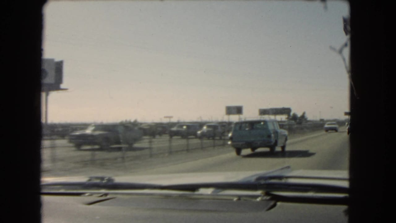 Late-frame windshield view of a 1966 California highway, with vintage cars ahead, distant roadside signs, median posts, dashboard trim, and faded 8mm color.