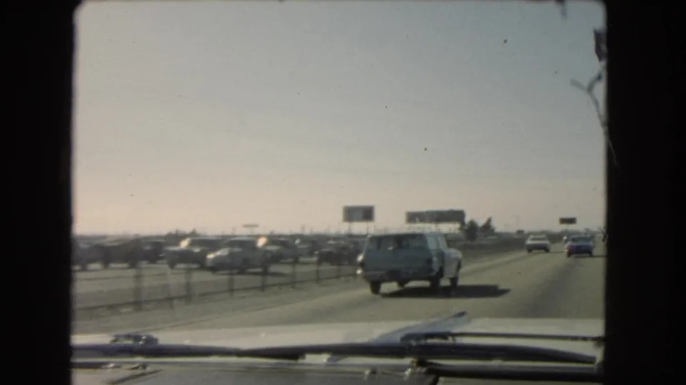 1966 California freeway traffic filmed from inside a vehicle, with a light station wagon ahead and billboards in the distance.