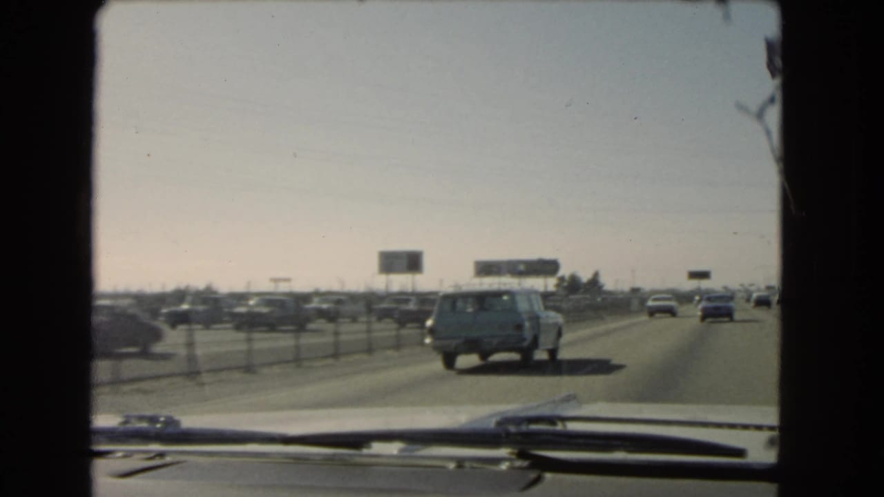 Interior vehicle view of 1966 California freeway traffic, with cars spread across lanes and low billboards on the horizon.