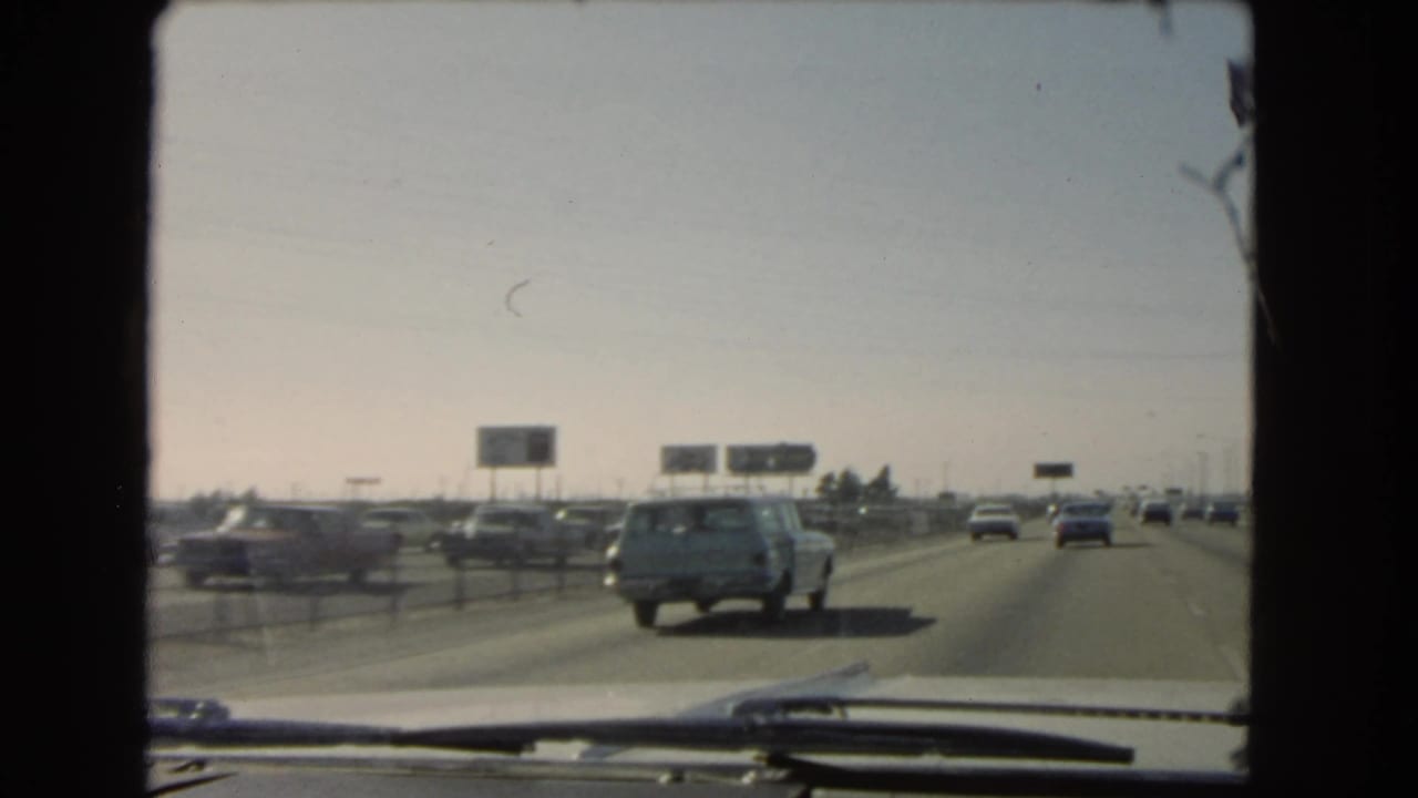 A light station wagon leads the windshield view as traffic continues along a California freeway in the supplied 1966 home-movie footage.