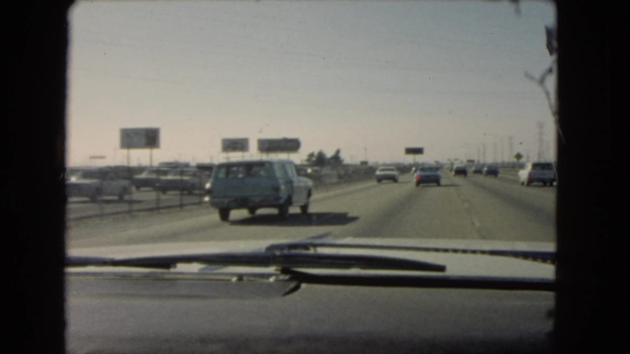 Late-frame windshield view of a 1966 California freeway, showing multiple cars, roadside signs, and the vehicle dashboard in the foreground.