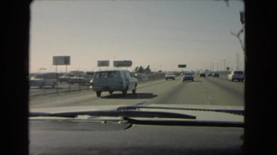 Late-frame windshield view of a 1966 California freeway, showing multiple cars, roadside signs, and the vehicle dashboard in the foreground.