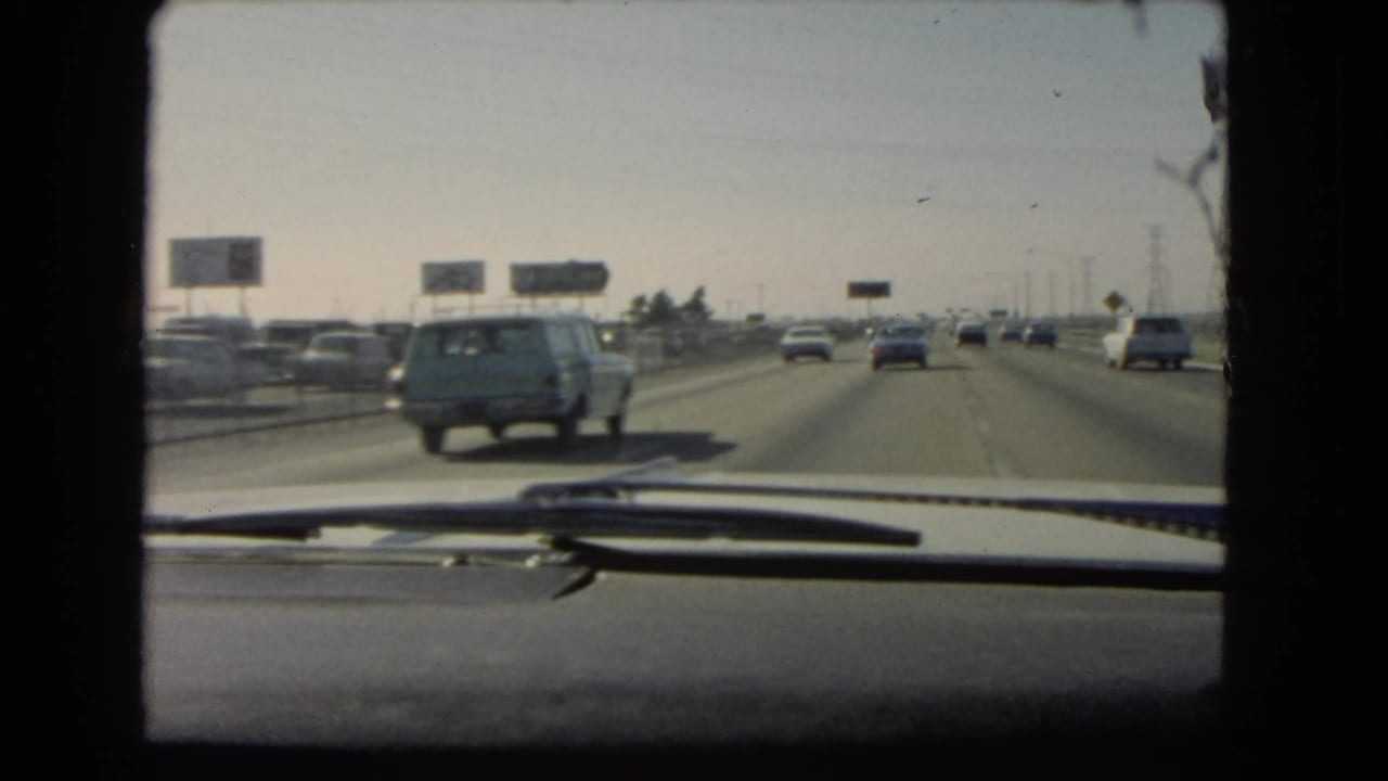Windshield view of a busy freeway with 1960s cars and a blue station wagon ahead.