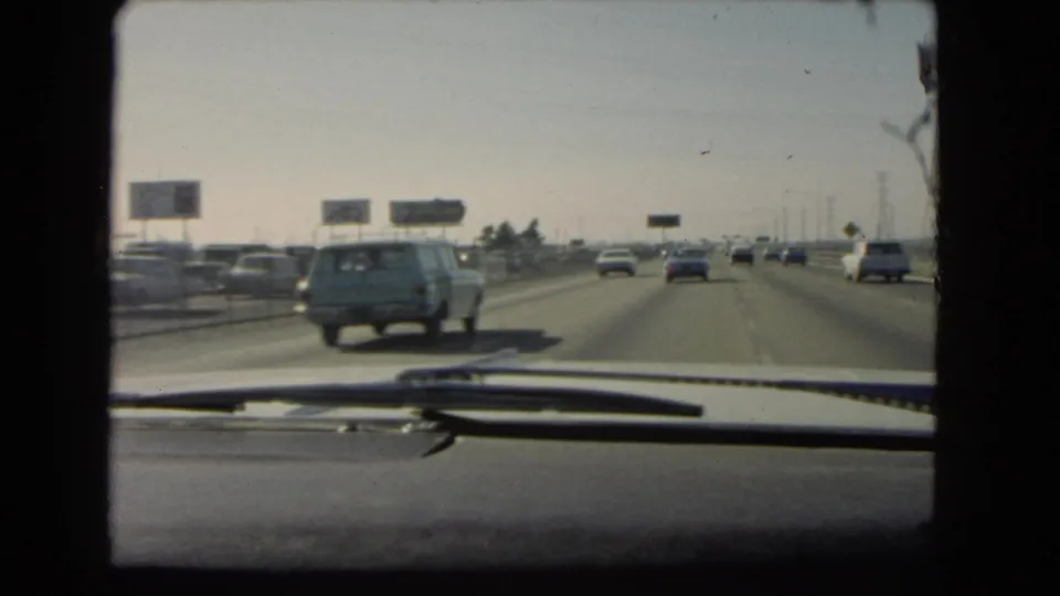 1966 California freeway scene filmed through a car windshield, with traffic, dashboard edge, and a blue station wagon in the lane ahead.