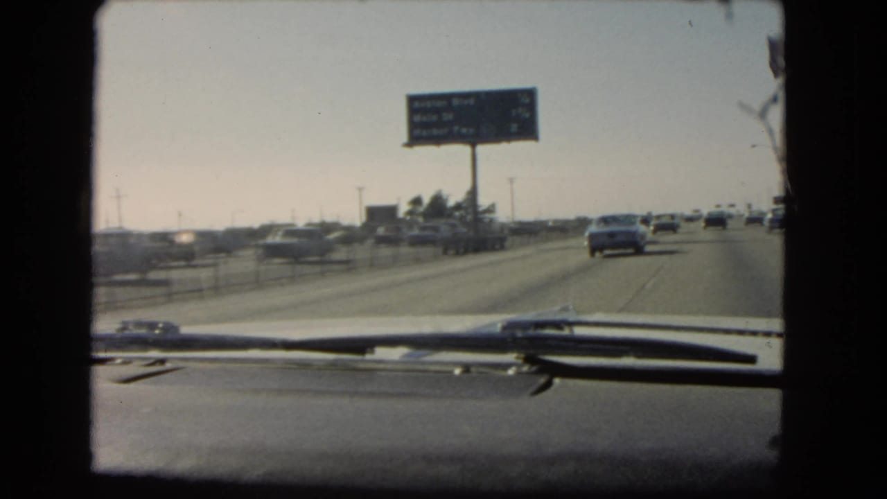 Windshield view on a 1966 California freeway with cars ahead, a roadside sign, dashboard trim, and windshield wipers visible.