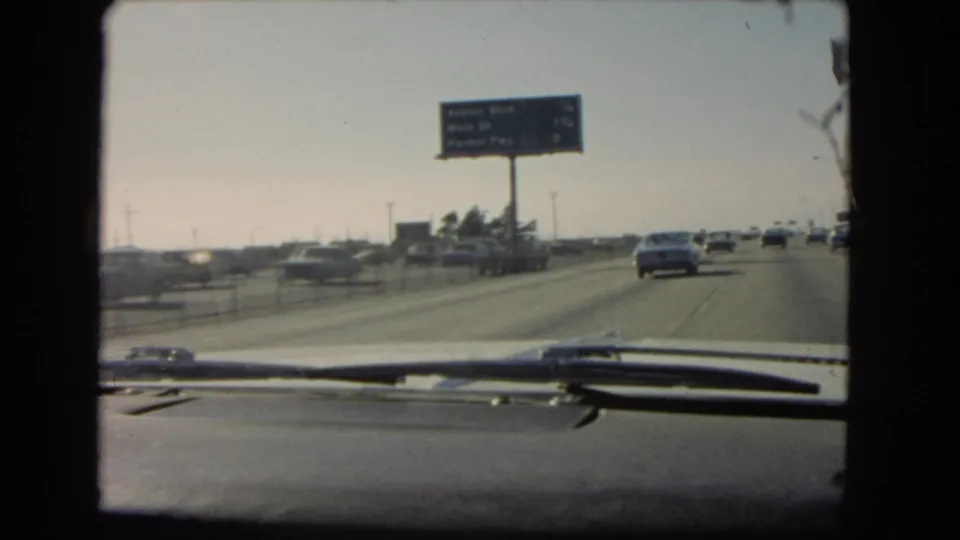 Windshield view on a 1966 California freeway with cars ahead, a roadside sign, dashboard trim, and windshield wipers visible.