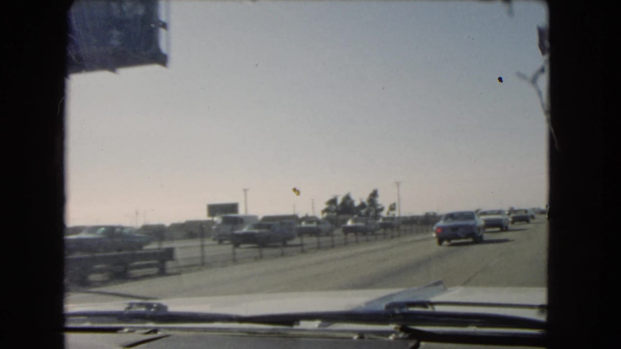 End of a 1966 California freeway driving shot, with cars ahead, a low roadside sign, windshield wipers, and bright hazy sky.