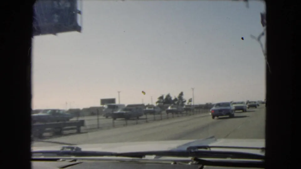 End of a 1966 California freeway driving shot, with cars ahead, a low roadside sign, windshield wipers, and bright hazy sky.