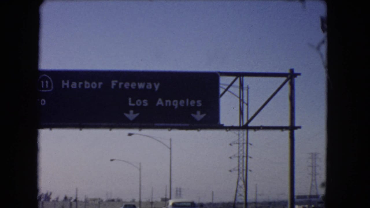 1966 California home-movie frame of a Harbor Freeway sign directing traffic toward Los Angeles, with roadway infrastructure beyond.