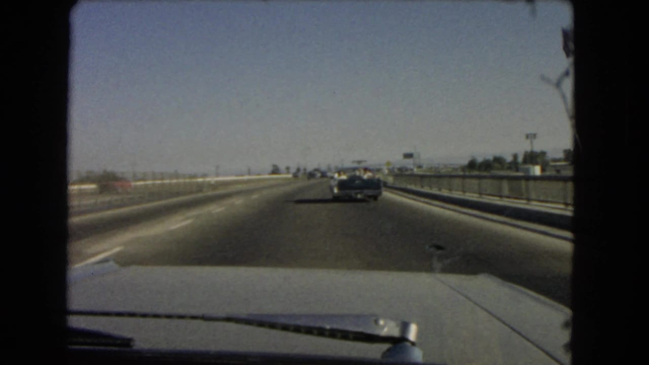 Driver-perspective 1966 California freeway shot with a car hood in the foreground and traffic moving ahead under clear sky.