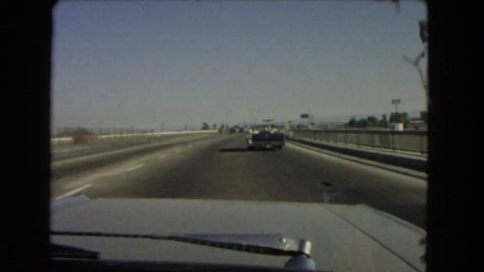 Driver-perspective 1966 California freeway shot with a car hood in the foreground and traffic moving ahead under clear sky.