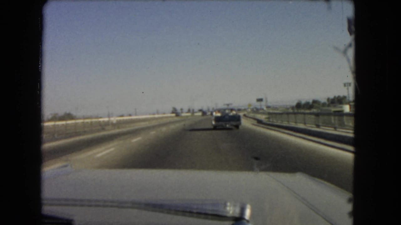1966 California highway travel seen through a windshield, with open lanes, a car ahead, and guardrails along the roadway.