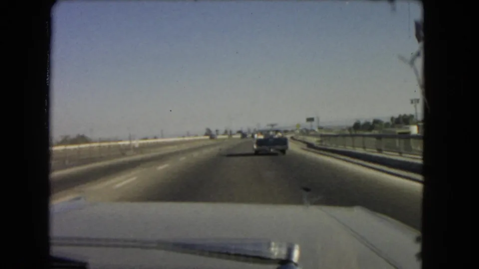 1966 California highway travel seen through a windshield, with open lanes, a car ahead, and guardrails along the roadway.