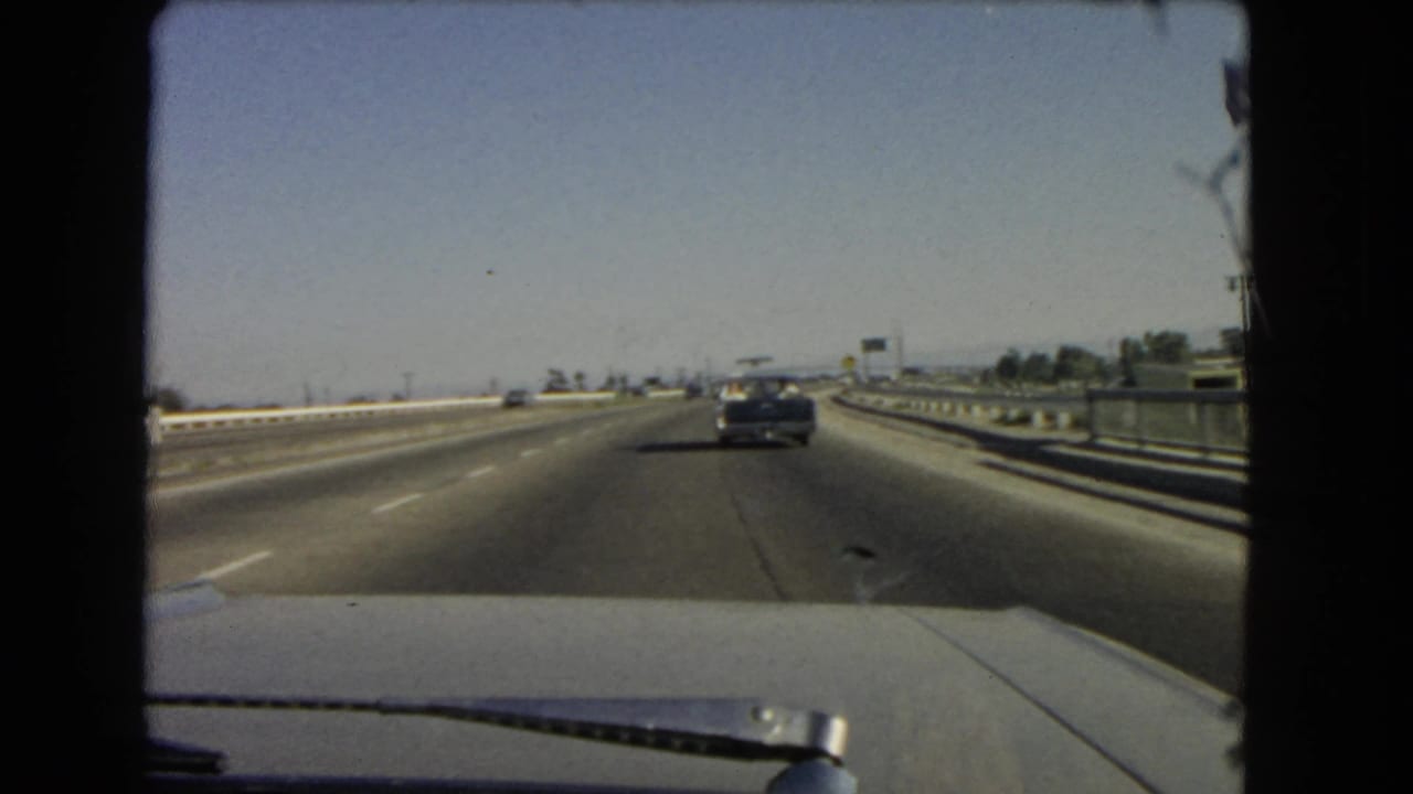 Late clip frame from a 1966 California freeway drive, showing a gentle road curve, guardrails, and a car ahead through the windshield.
