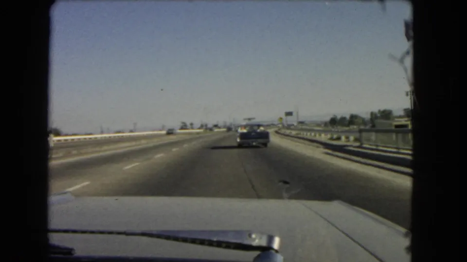 Late clip frame from a 1966 California freeway drive, showing a gentle road curve, guardrails, and a car ahead through the windshield.