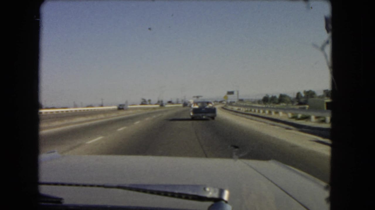 1966 California 8mm windshield view of a freeway with light traffic, a dark sedan ahead, guardrails, and clear sky.