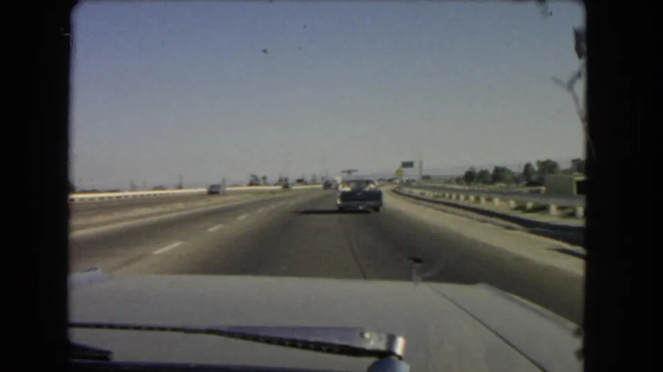 1966 California 8mm windshield view of a freeway with light traffic, a dark sedan ahead, guardrails, and clear sky.