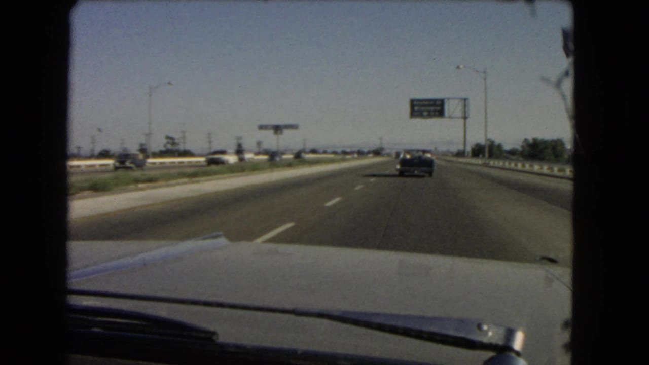 Freeway lanes viewed over a car hood with a dark sedan ahead and an overhead green sign at right.