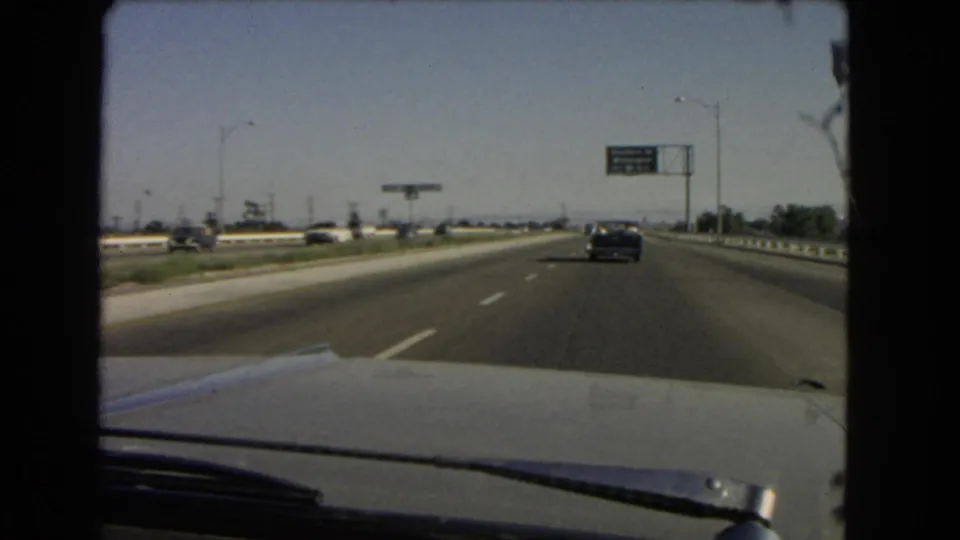 1966 California highway scene from inside a moving car, with a dark sedan ahead and a green overhead road sign approaching.