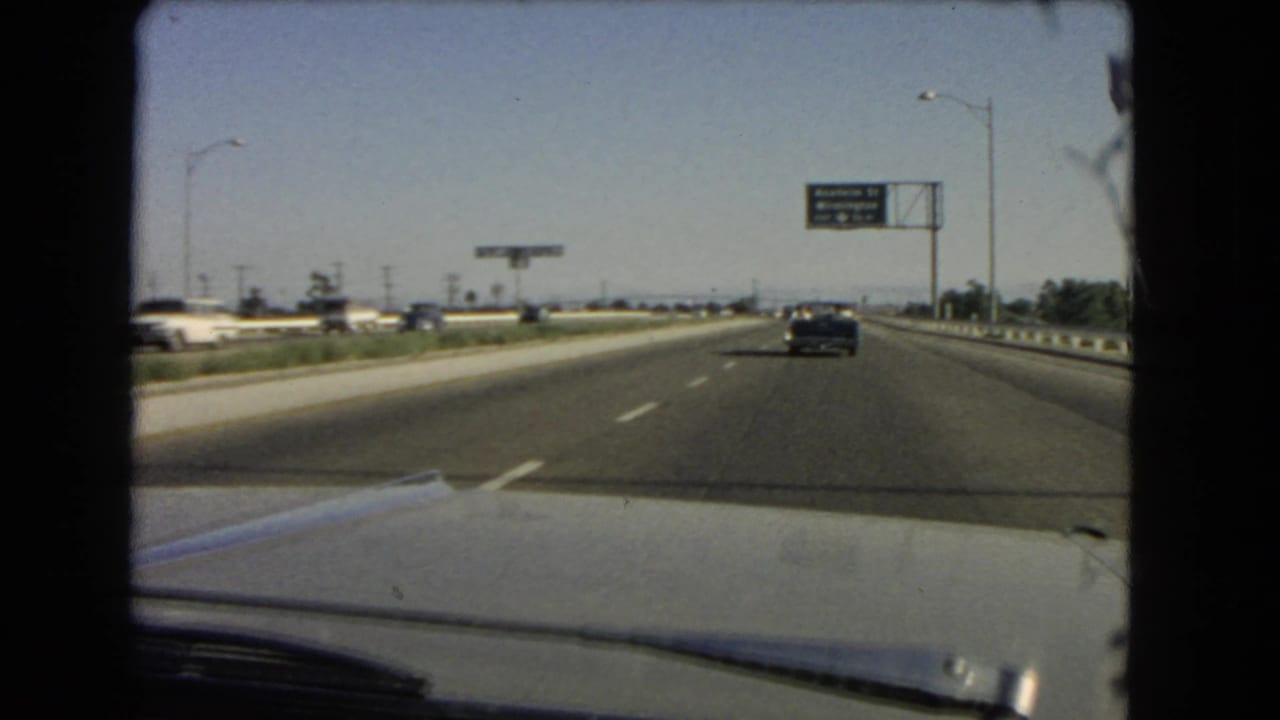 Mid-1960s California freeway footage shows a windshield view past the car hood toward a dark sedan and overhead highway signage.