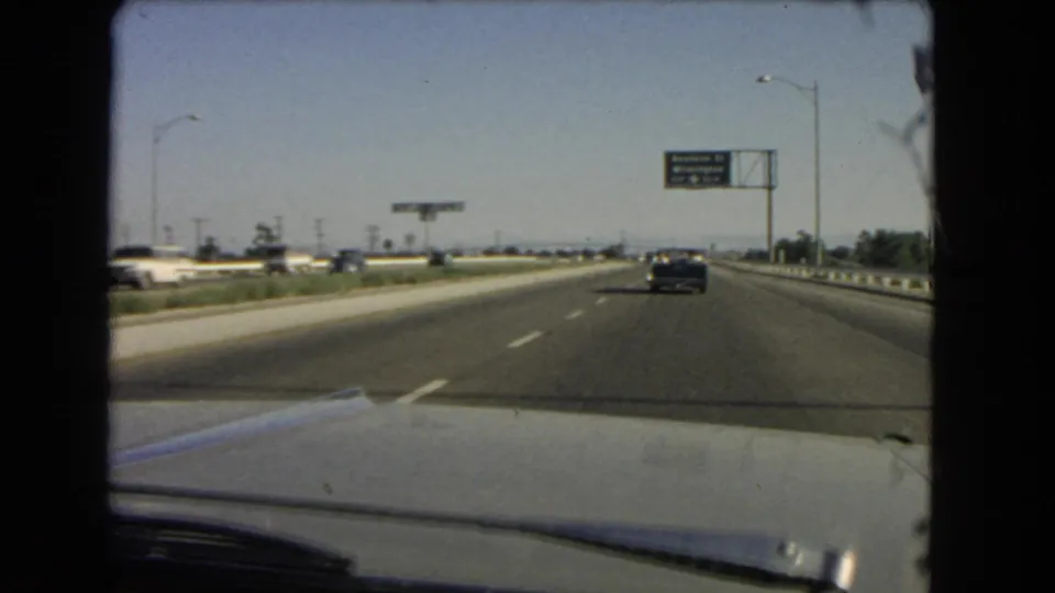 Mid-1960s California freeway footage shows a windshield view past the car hood toward a dark sedan and overhead highway signage.