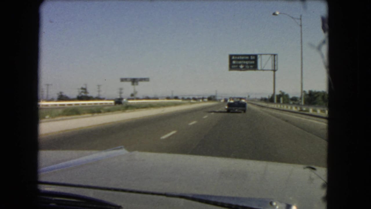 1966 California 8mm frame of freeway driving, with the car hood in foreground, a dark sedan ahead, and green directional signage at right.