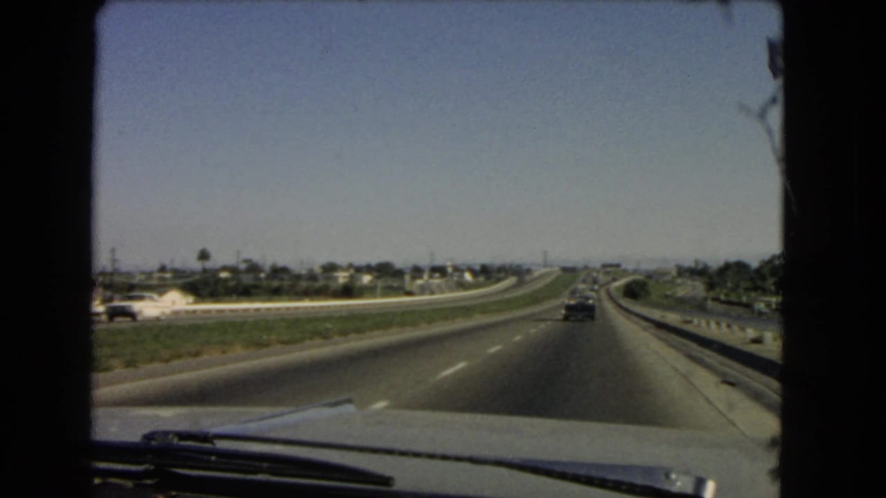 1966 California home-movie view through a windshield along an interstate, with light traffic, distant houses, palm trees, and road barriers.