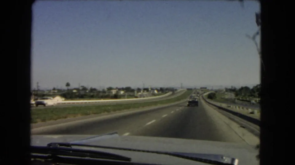 1966 California home-movie view through a windshield along an interstate, with light traffic, distant houses, palm trees, and road barriers.