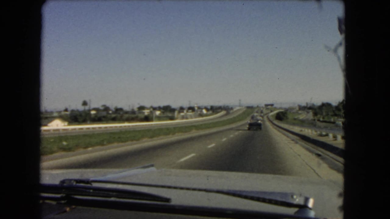 1966 California interstate scene filmed from inside a moving car, showing a curved roadway, light traffic, suburban houses, and utility poles.