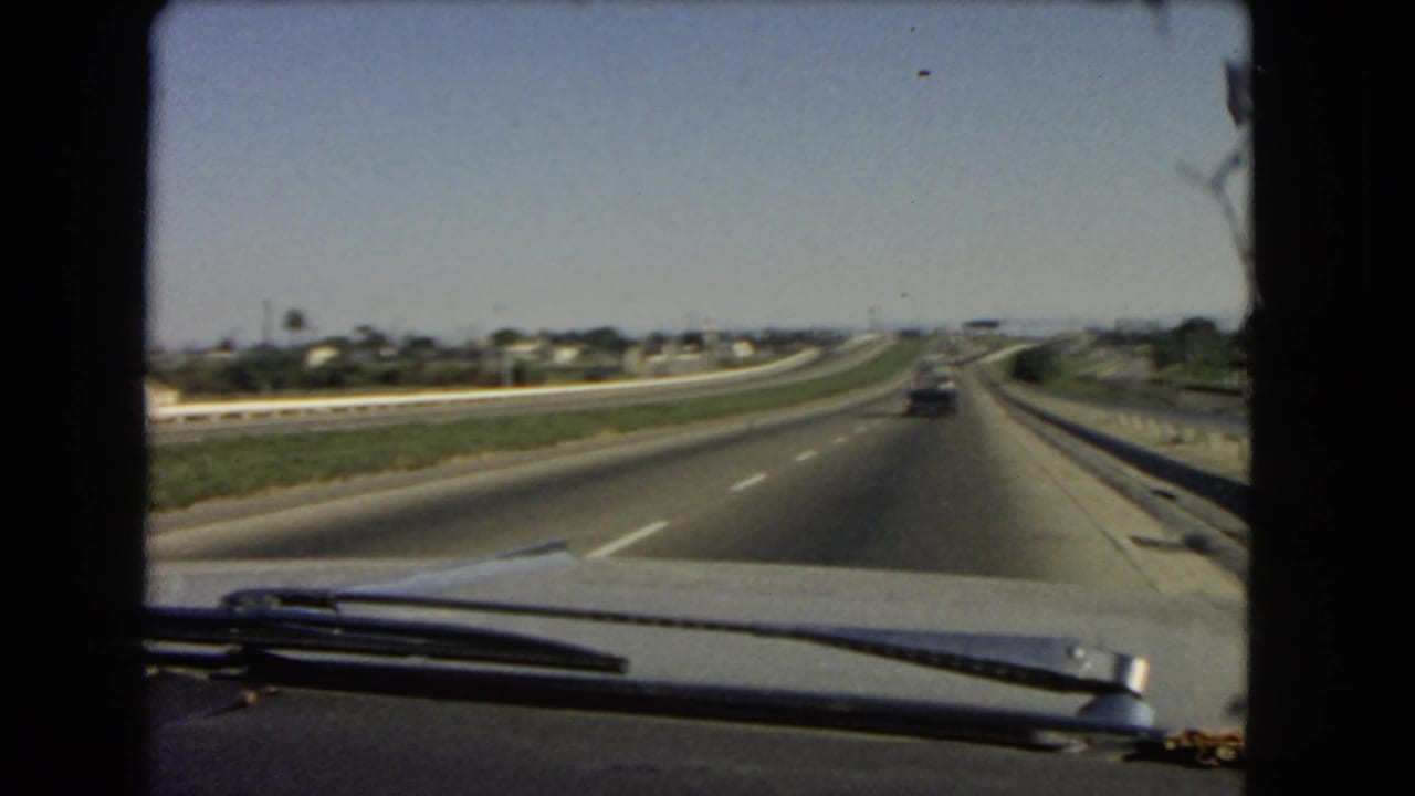 Archival 8mm windshield footage from 1966 California, showing a straight interstate view with light traffic and low suburban buildings in the distance.