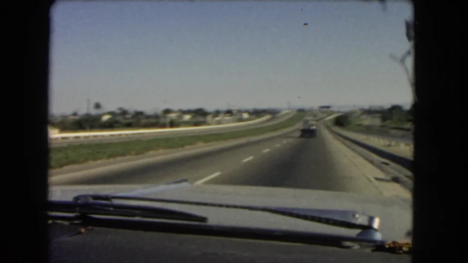 Archival 8mm windshield footage from 1966 California, showing a straight interstate view with light traffic and low suburban buildings in the distance.