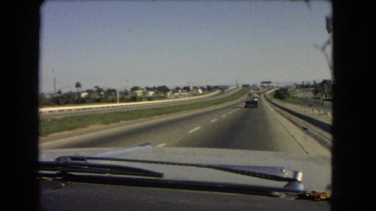 1966 California interstate footage from a moving car, with the hood and wipers in the foreground and suburban roadside features beyond the lanes.