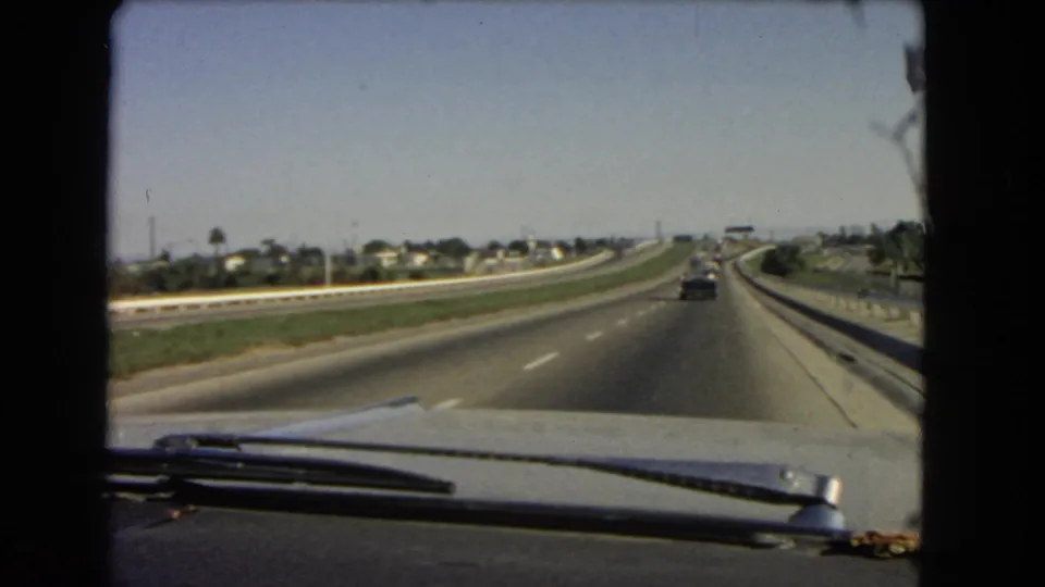 1966 California interstate footage from a moving car, with the hood and wipers in the foreground and suburban roadside features beyond the lanes.