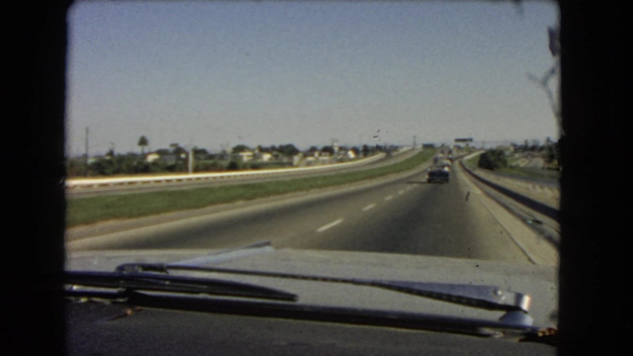 1966 California home-movie frame looking through a car windshield at a divided highway, grassy median, distant houses, and cars ahead.