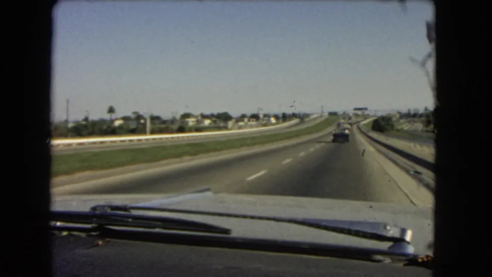 1966 California home-movie frame looking through a car windshield at a divided highway, grassy median, distant houses, and cars ahead.
